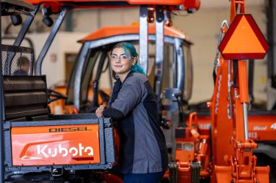 A Chemeketa Diesel Technology student with blue hair is in the garage lab surrounded by Kubota equipment. She is leaning against the tailgate of a four-wheel buggy.